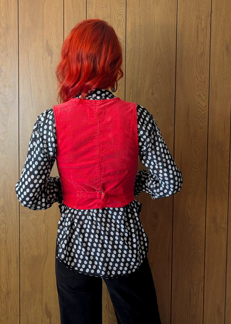 Person wearing a red vest over a black and white polka dot shirt against a wooden wall.