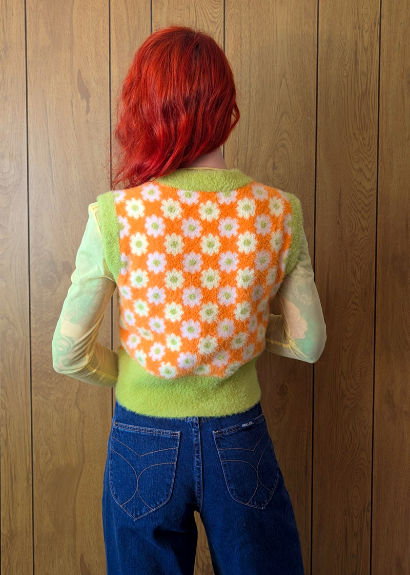 Person wearing a green sweater vest  with an orange floral pattern on the back, standing against a wooden wall.