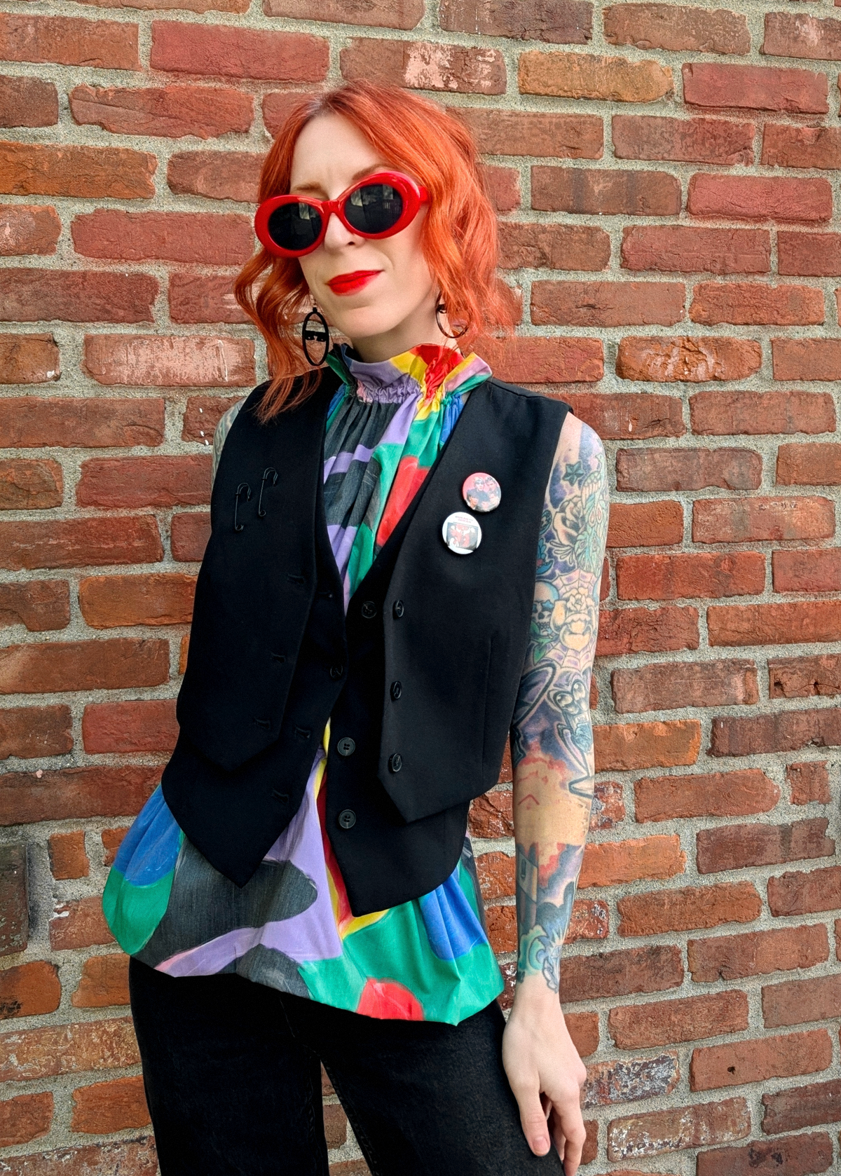 Person with red hair wearing a colorful halter top with a black vest buttoned over top, paired with black jeans, standing against a brick wall