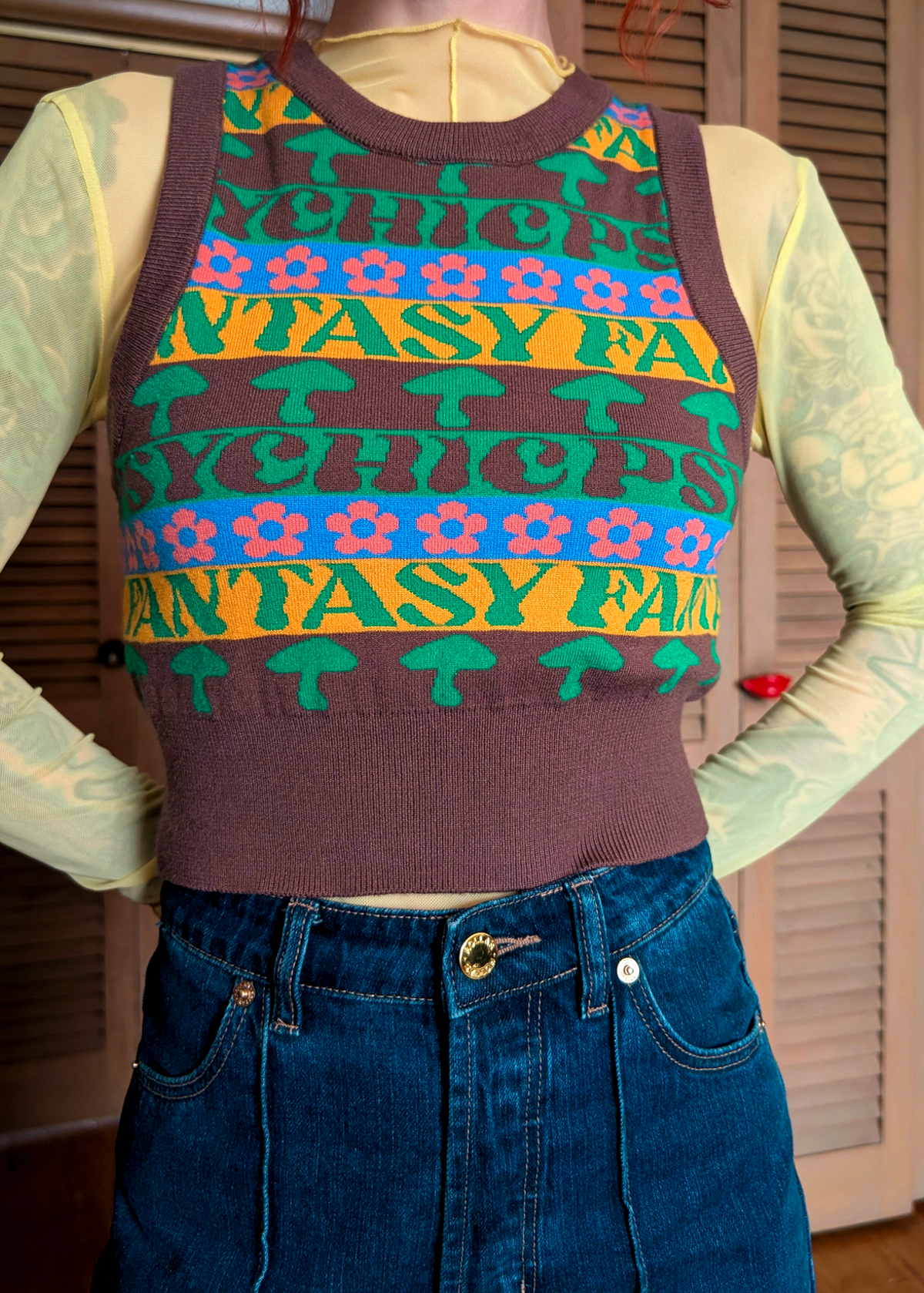 Person wearing a colorful patterned sweater vest over a light yellow mesh shirt and jeans