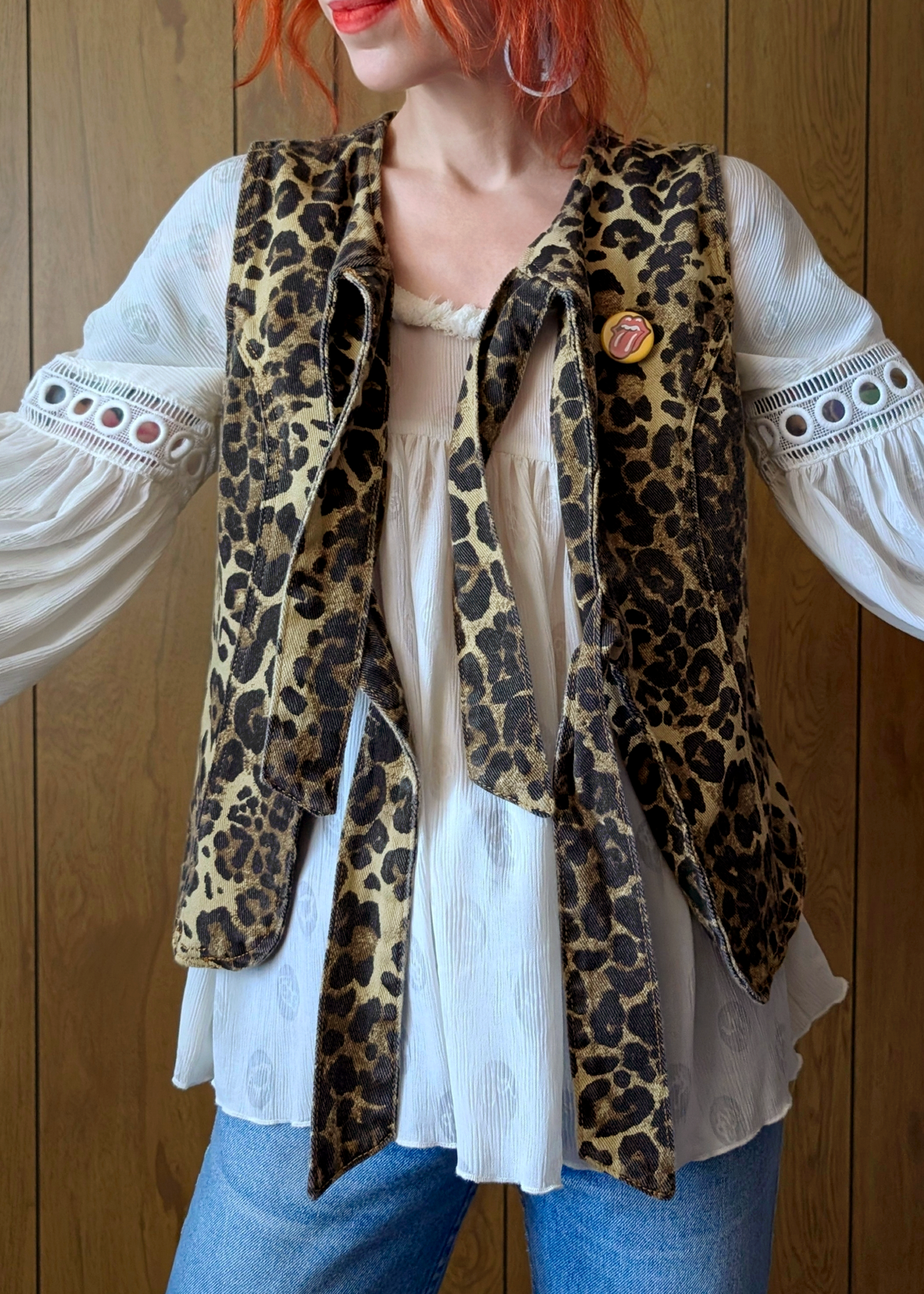 Person wearing a leopard print vest over a white blouse with blue jeans against a wooden background