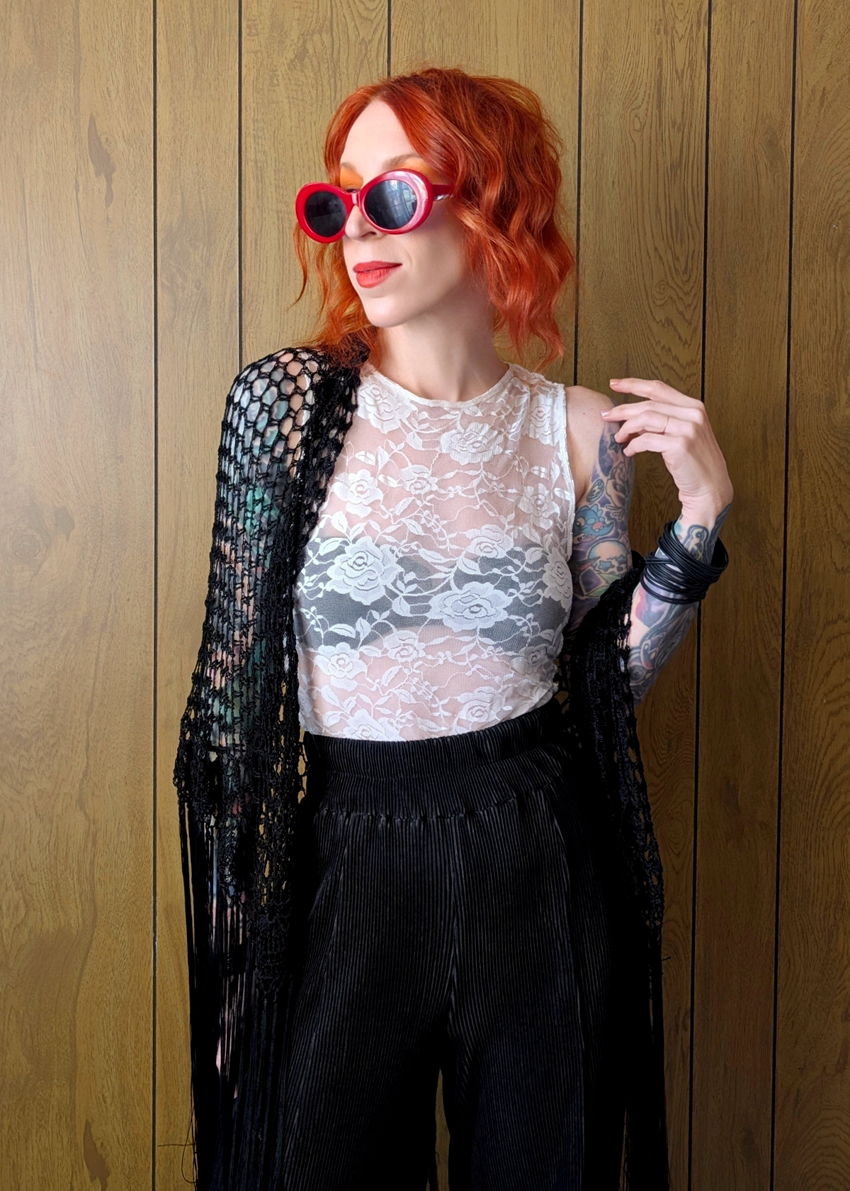 Person with red hair wearing sunglasses, a lace top, and black pants against a wooden wall.