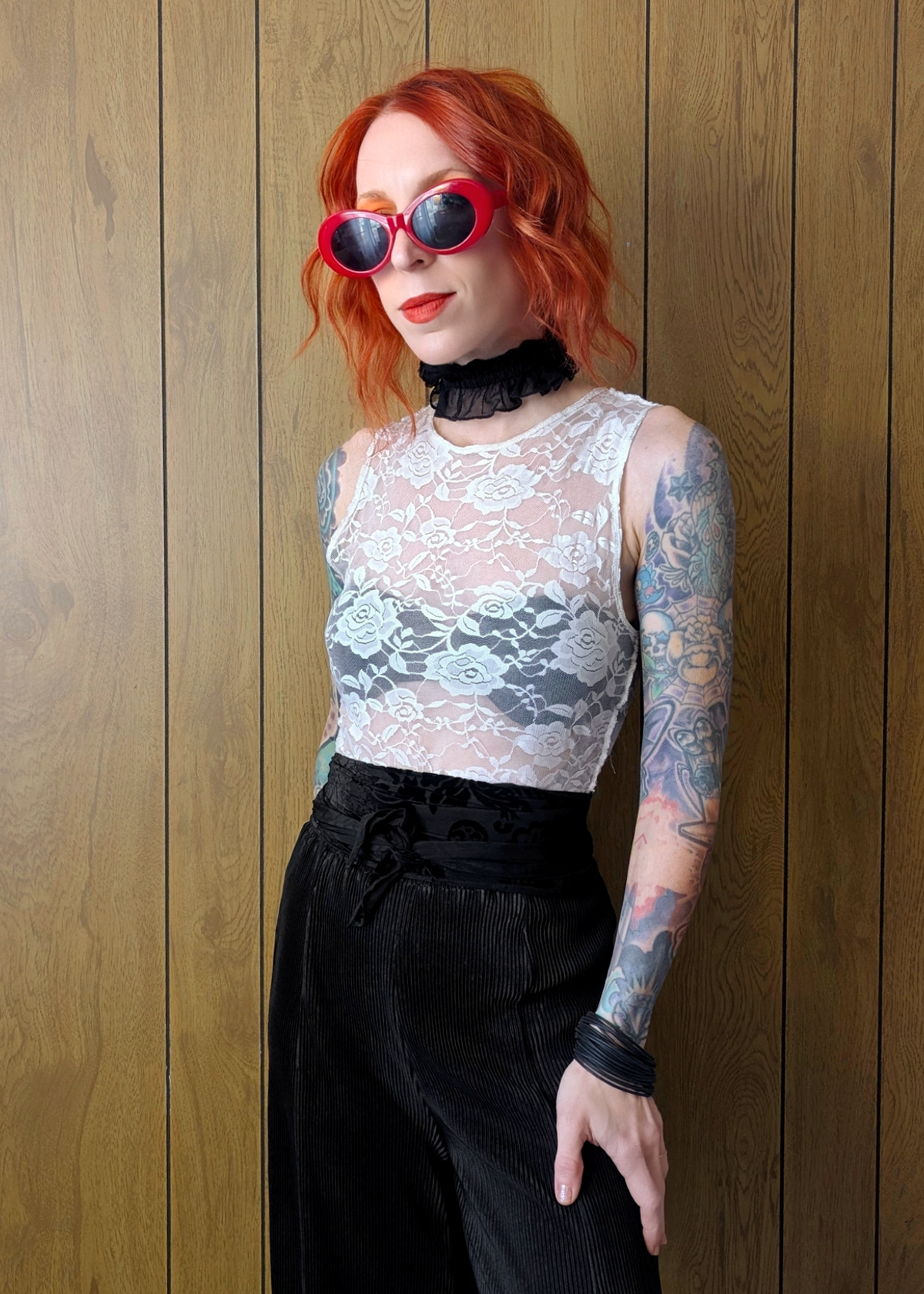 Person with red hair wearing a white lace sleeveless top and black pants against a wooden panel background