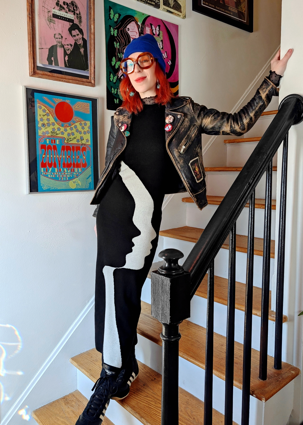 Person standing on a staircase wearing a black and white knit dress with a face design, styled with a leather jacket, surrounded by colorful artwork on the wall.