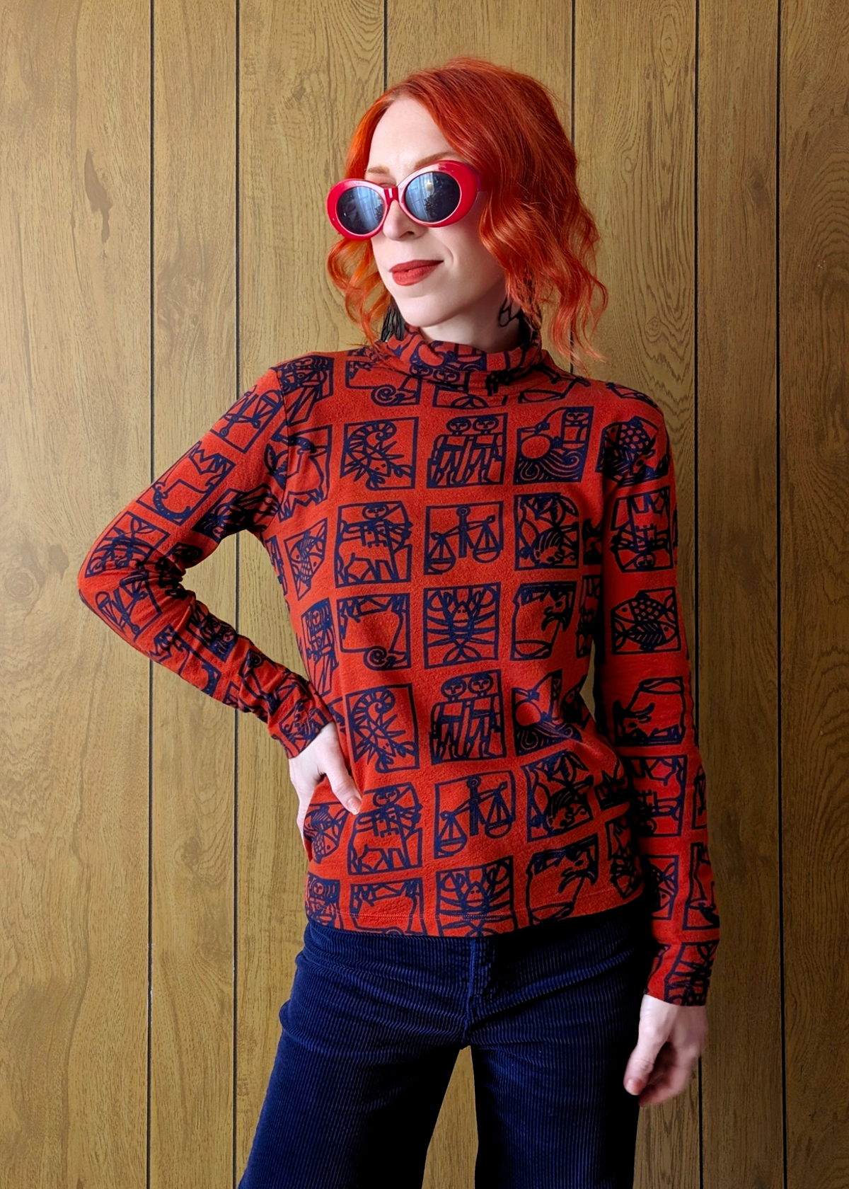 Person wearing a red zodiac-patterned long-sleeve top and navy corduroys against a wooden wall.
