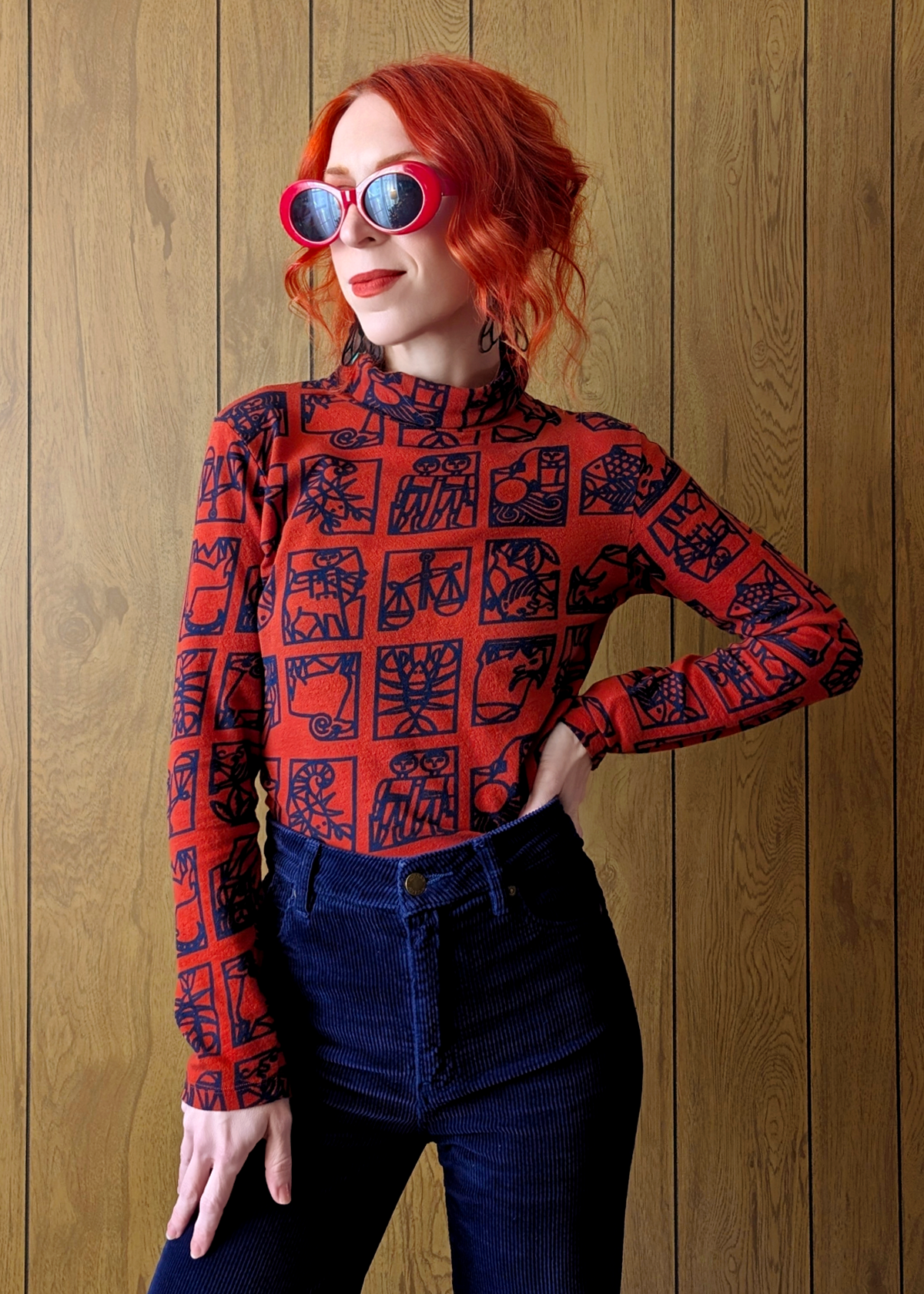 Person wearing a red zodiac-patterned long-sleeve top and navy corduroys against a wooden wall.